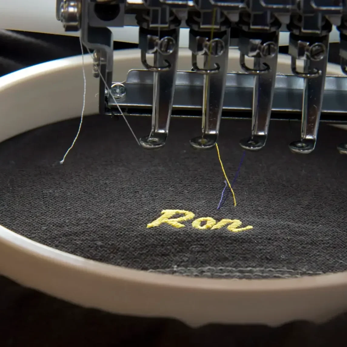 Embroidery machine stitching the name 'Ron' in yellow thread on black fabric.